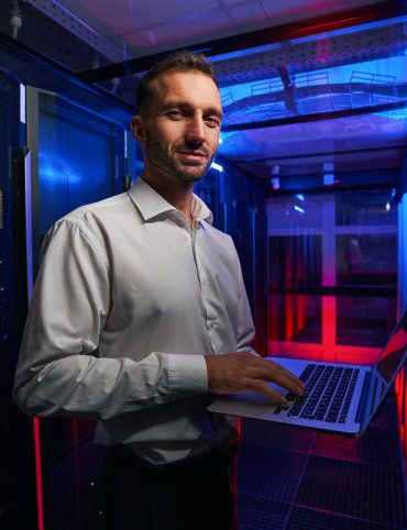 Bearded smiling system administrator is working with server rack and telecommunication equipments in